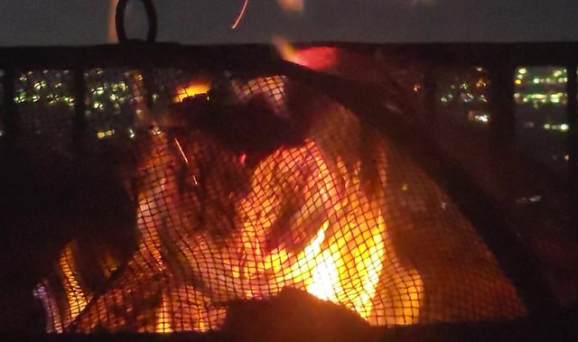 A fire burning under a mesh spark screen at dusk, city lights in the valley behind a railing